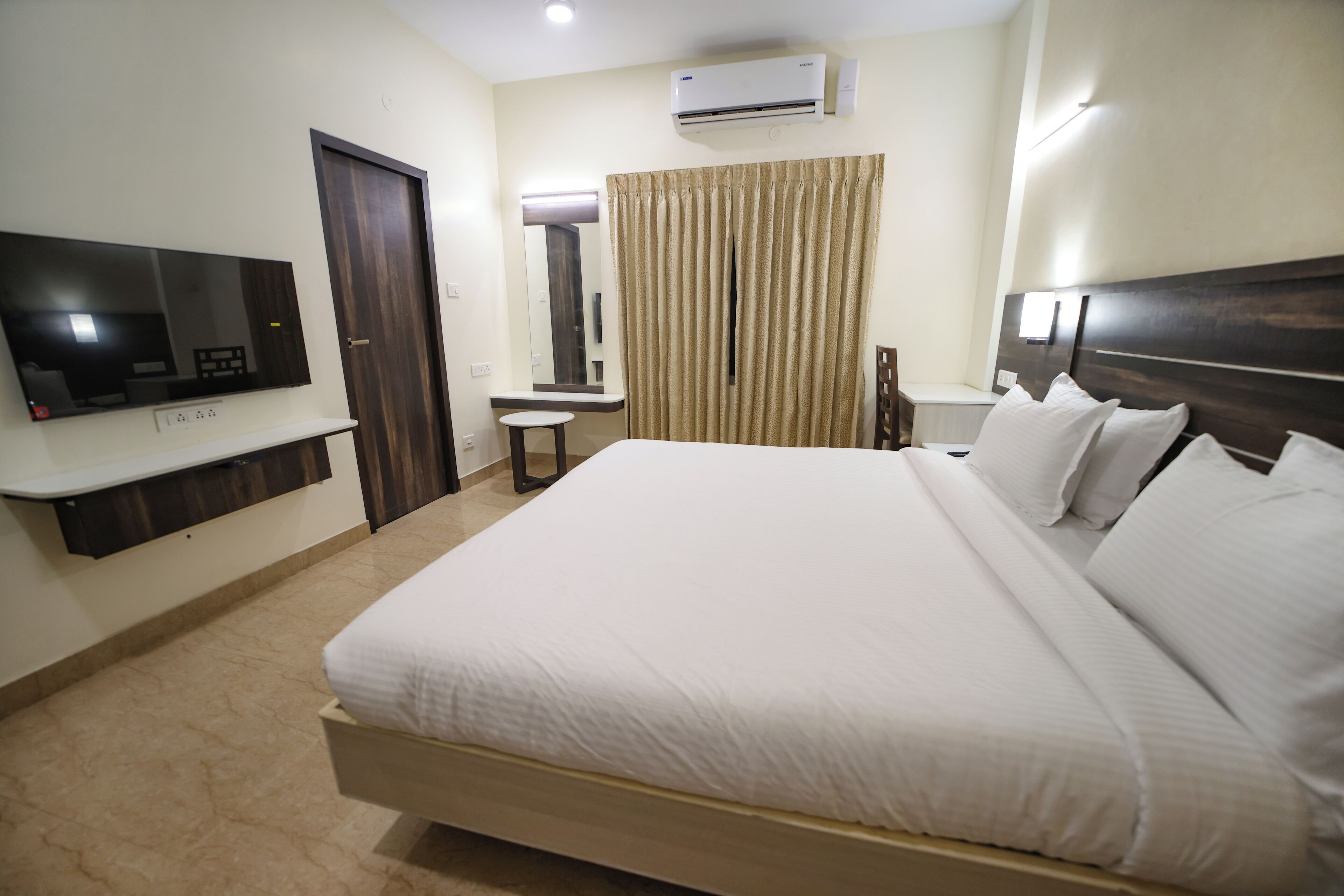 A hotel room with a large bed, a flat-screen TV mounted on the wall, a small table, a mirror, and a window with curtains.