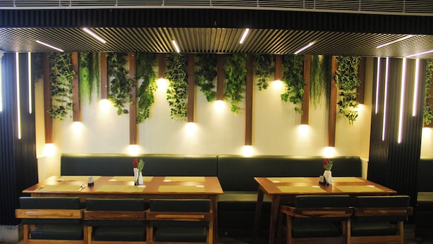 A restaurant interior with wooden tables and benches, a green cushioned bench, and hanging plants. - Hotel Pavitra (Purnia)