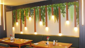 A restaurant with wooden tables and green upholstered booths, featuring hanging plants and wall-mounted lights. - Hotel Pavitra (Purnia)