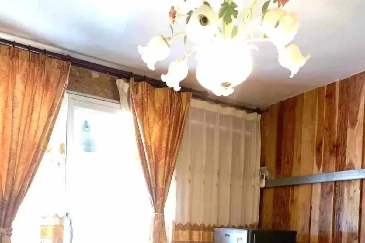 Quarto com cama, cortinas, ventoinha no teto e um pequeno frigorífico.