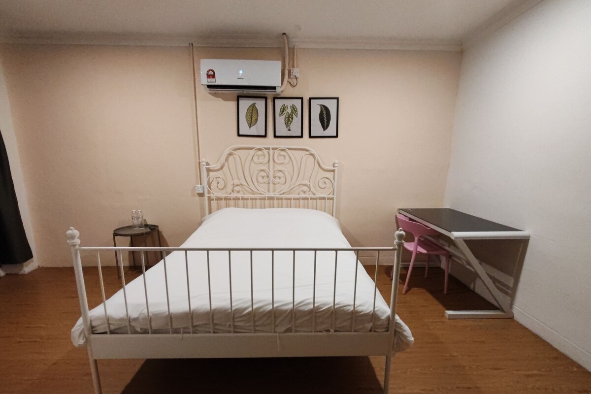 A bedroom with a white metal bed, a small bedside table, a desk with a chair, and an air conditioner on the wall.