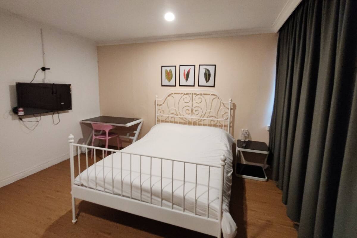 A bedroom with a bed, a desk, a chair, a TV mounted on the wall, and a window with curtains.