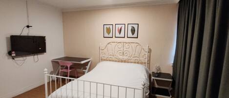 A bedroom with a white metal bed, a small desk with a chair, a television mounted on the wall, and a window with curtains.