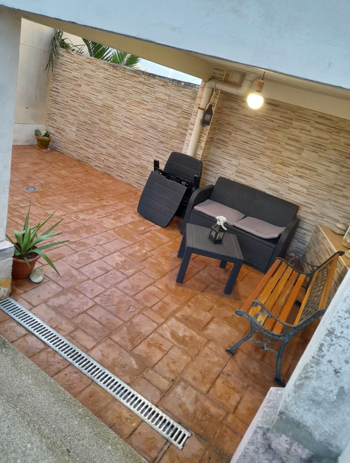 A patio with a sofa, a chair, and a table. There are potted plants and a drainpipe.