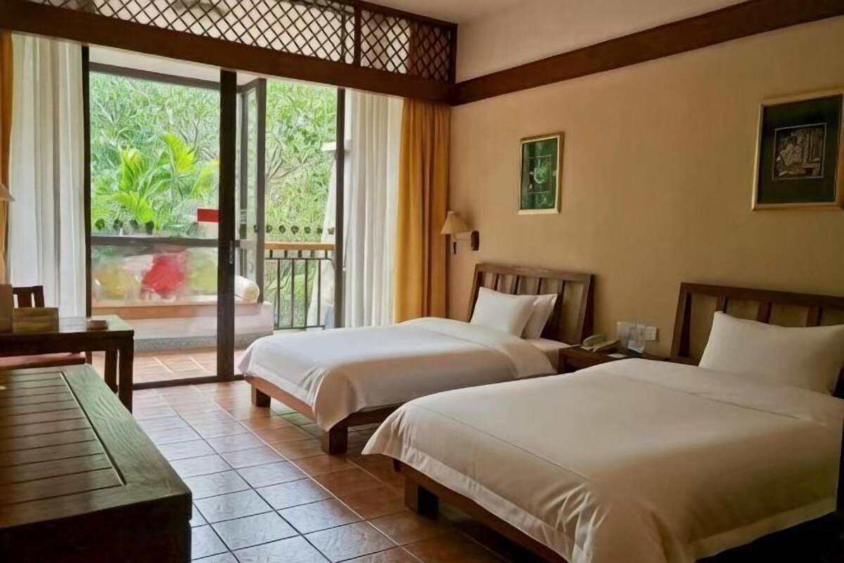 A hotel room with two beds, a wooden desk, a balcony with a view of greenery, and a door leading outside.