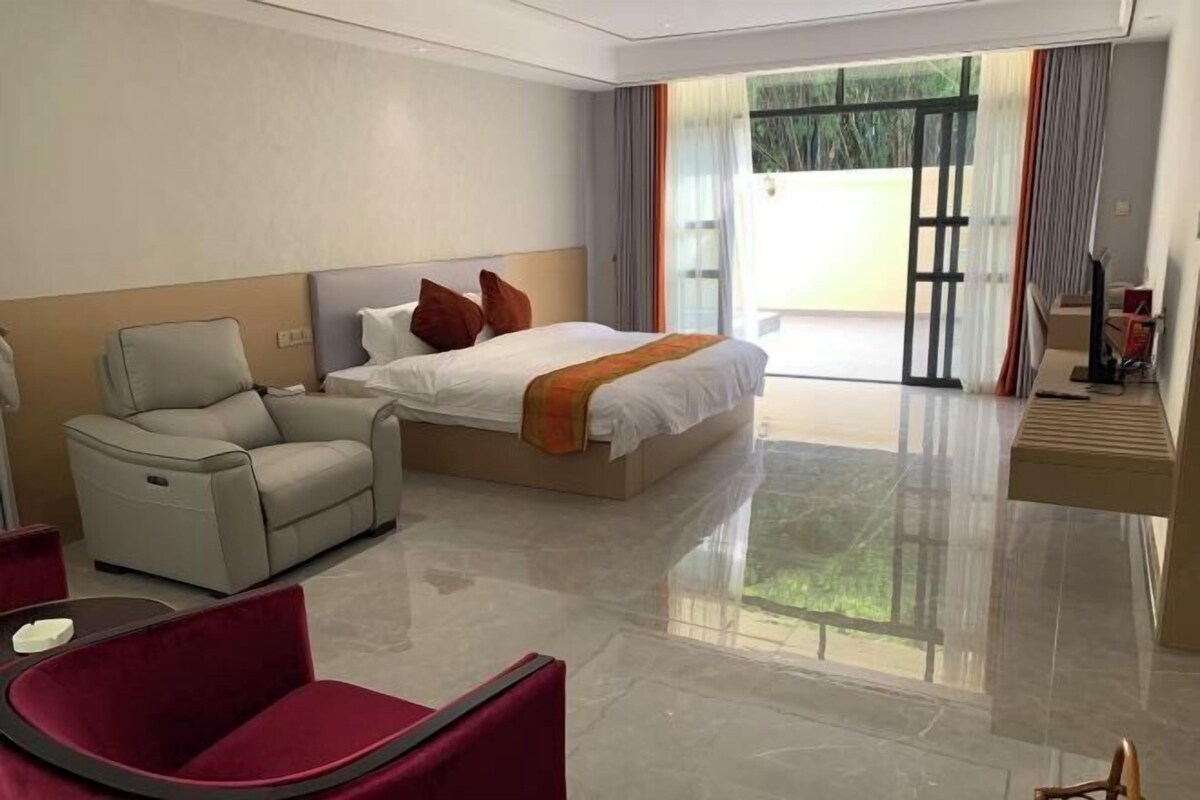 A modern hotel room with a large bed, a sofa, a chair, and a view of the outdoors through a glass door.