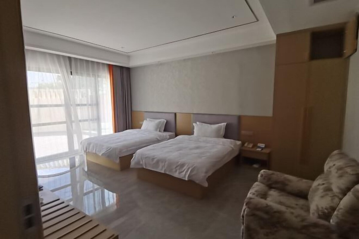 A hotel room with two beds, a wooden bench, a sofa, and a large window with sheer curtains.