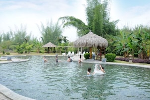 Una piscina con gente, rodeada de vegetación tropical y construcciones con techo de paja.