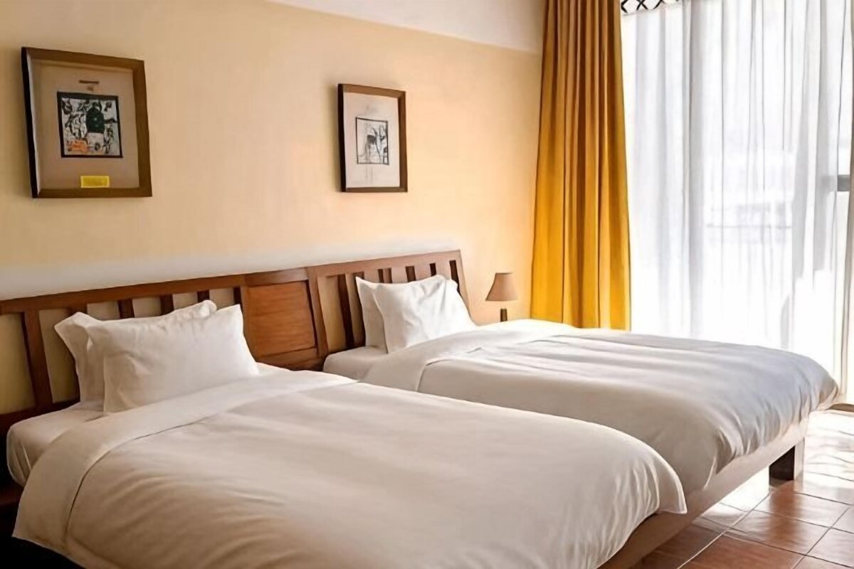 A hotel room with two beds, a wooden headboard, a nightstand, and a window with curtains.