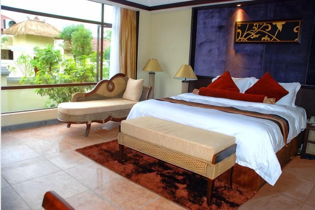 A hotel room with a large bed, a sofa, a window with a view of greenery, and a decorative wall piece.