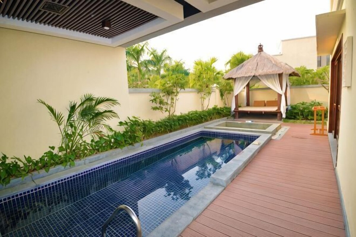 A swimming pool with a wooden deck, a thatched-roof gazebo, and lush greenery.