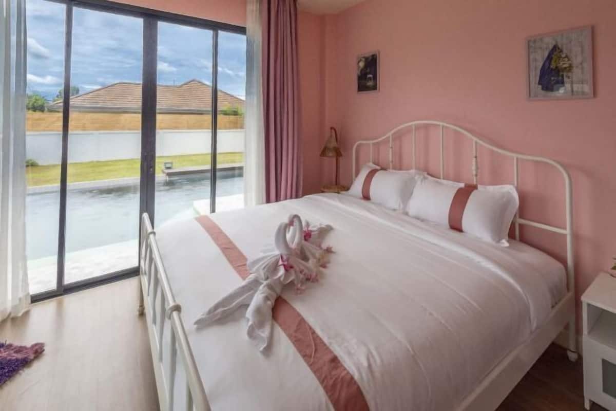 A bedroom with a bed, a nightstand, a window with a view of a house and a pool, and a pink wall.
