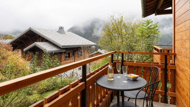 Un balcon en bois, agrémenté d’une table et de chaises, qui offre une vue sur une région montagneuse parsemée de maisons et d’arbres.