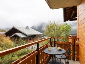 Un balcón de madera con una mesa y sillas, con vista a una zona montañosa con casas y árboles.