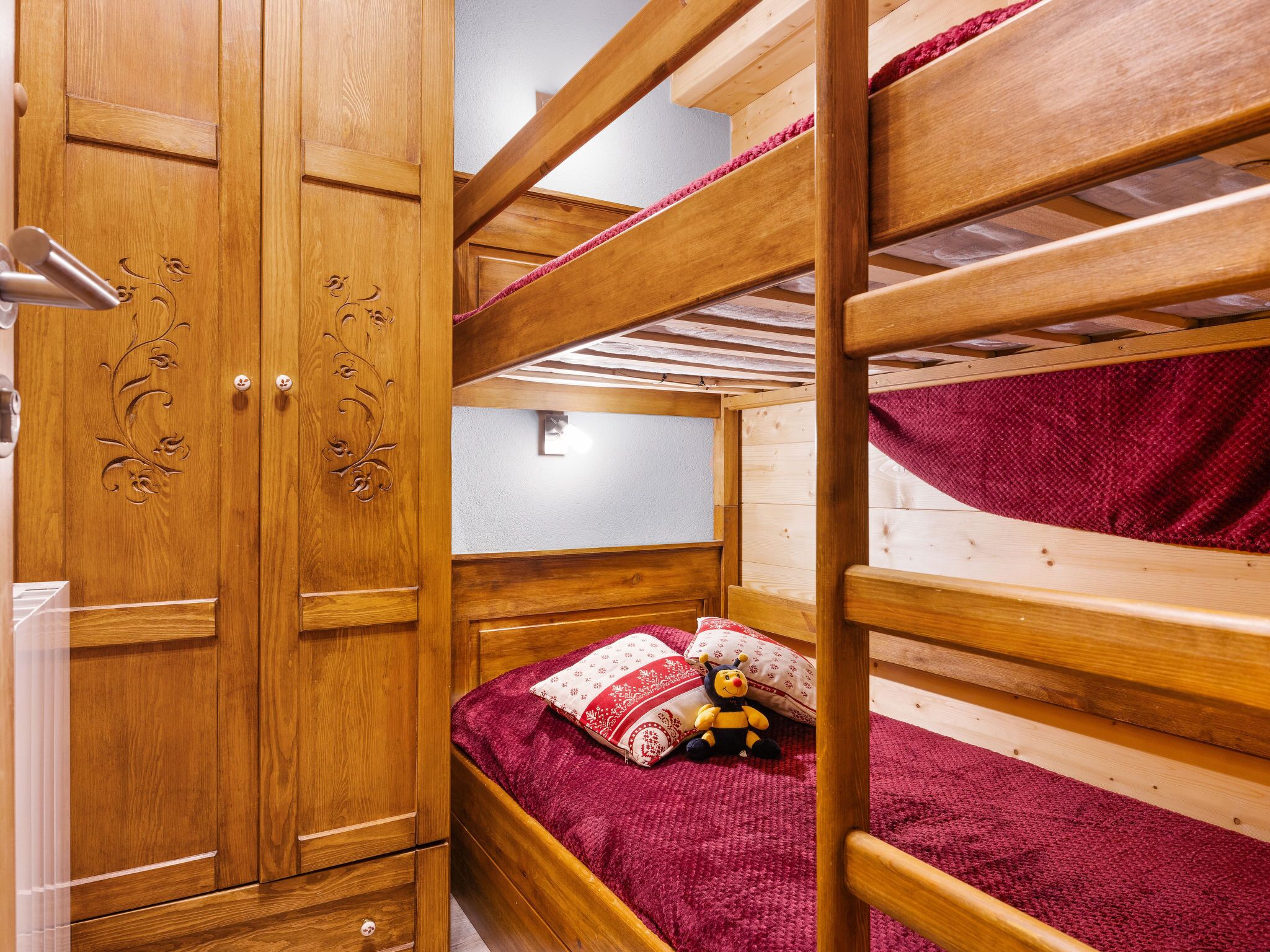Een slaapkamer met een stapelbed, een houten garderobe en een klein bed met een rode deken en kussens.