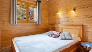 Une chambre à coucher dans une cabane en bois, avec un lit, des oreillers, une table de chevet, une fenêtre avec des rideaux et une petite table sur laquelle se trouve un livre.