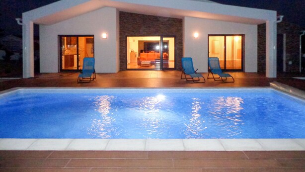 Una casa moderna con piscina, dos sillones de descanso y puerta de entrada de cristal.
