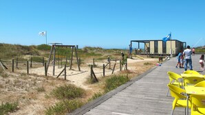 Un paseo marítimo en la playa con sillas amarillas, una pasarela de madera y un edificio con una bandera azul.