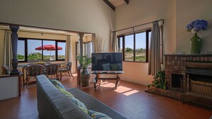 Amplia sala de estar con televisor grande, asientos cómodos, chimenea y ventanas amplias que ofrecen vistas del paisaje.