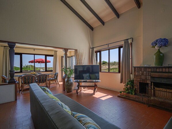 A spacious living room with a large TV, comfortable seating, a fireplace, and large windows offering a view of the landscape.