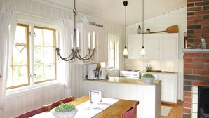 Et køkken med hvide skabe, et træspisebord med stole, en hængende lampe og en murstenspejs.