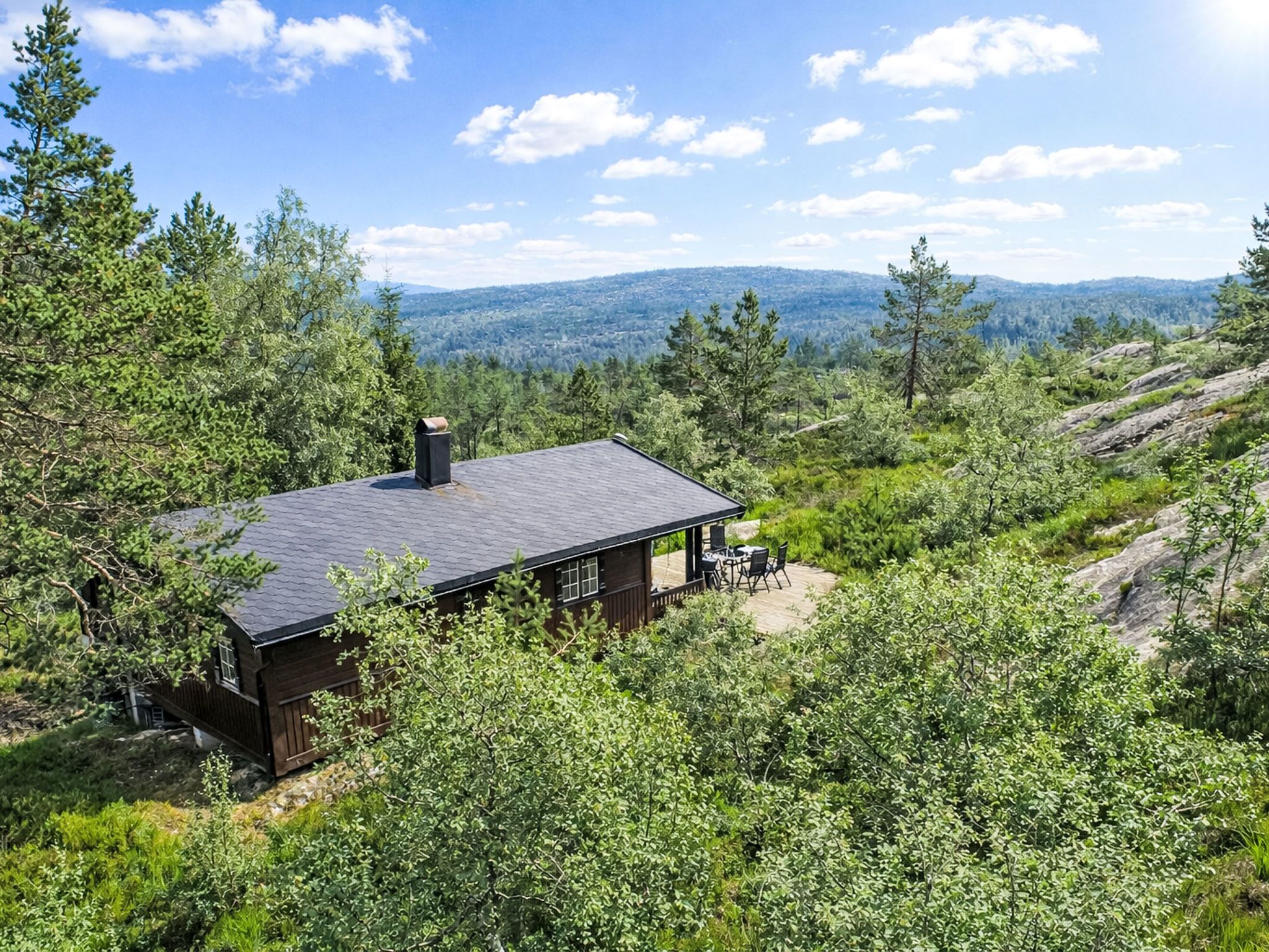 A cabin with a dark roof, surrounded by dense green foliage and rocky terrain, overlooking a distant mountain range.