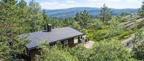 A cabin with a dark roof, surrounded by dense green foliage and rocky terrain, overlooking a distant mountain range.