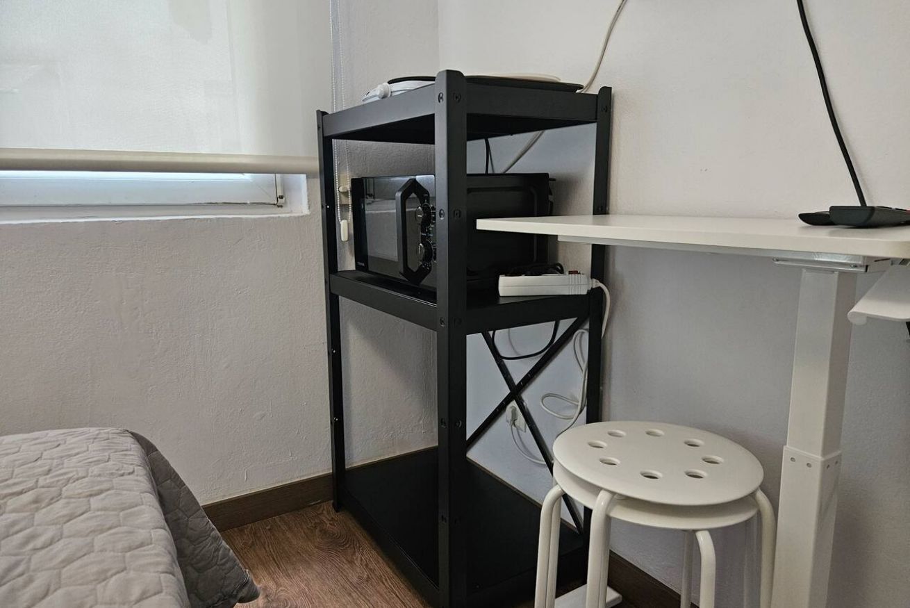 Habitación pequeña con cama, un taburete blanco, un escritorio de metal negro con una repisa blanca y una pared blanca.