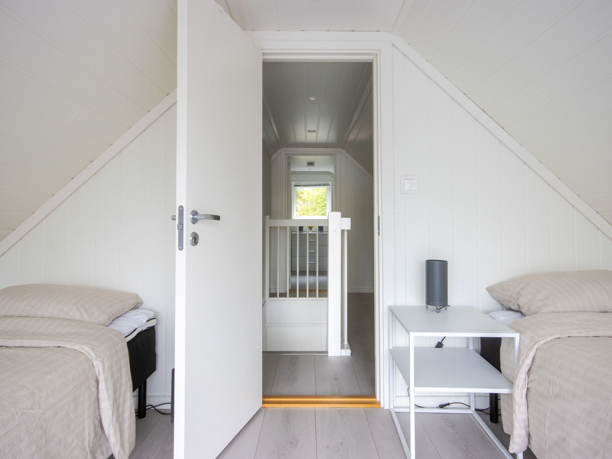 A small, white, attic-style room with two beds, a small table, and a door leading to a staircase.