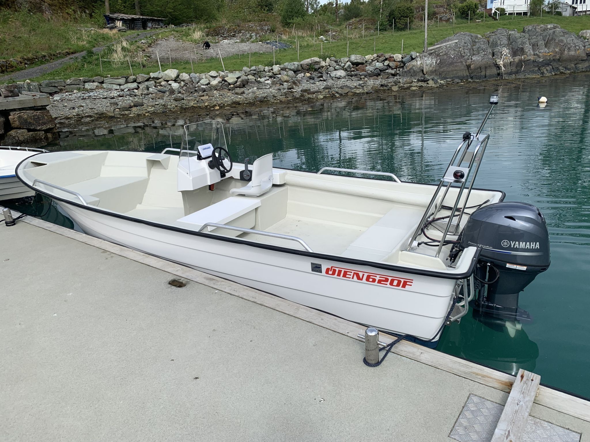 Een kleine, witte motorboot aangemeerd aan een betonnen steiger, met een rotsachtige kustlijn en een huis op de achtergrond.