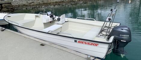 Een kleine, witte motorboot aangemeerd aan een betonnen steiger, met een rotsachtige kustlijn en een huis op de achtergrond.