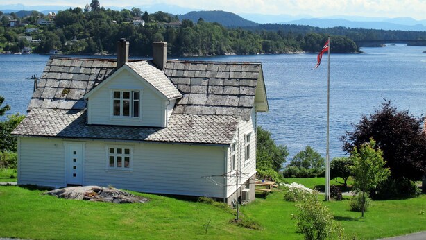 Ein weißes Haus mit einem Stehendach, einer Fahnenstange mit einer Flagge und im Hintergrund ein See und Berge.