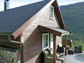 Una cabaña de madera con techo inclinado, un porche con mobiliario exterior y vistas a un paisaje montañoso.