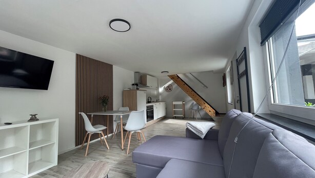 A modern living room with a gray sofa, a dining area with white chairs, a kitchen with wooden cabinets, and a staircase leading to an upper level. - Oase im Grünenhinterhof, EG, Ruhe, Terrasse, Carport, Free Paytv, Kaffee&tee, (Duisburg)