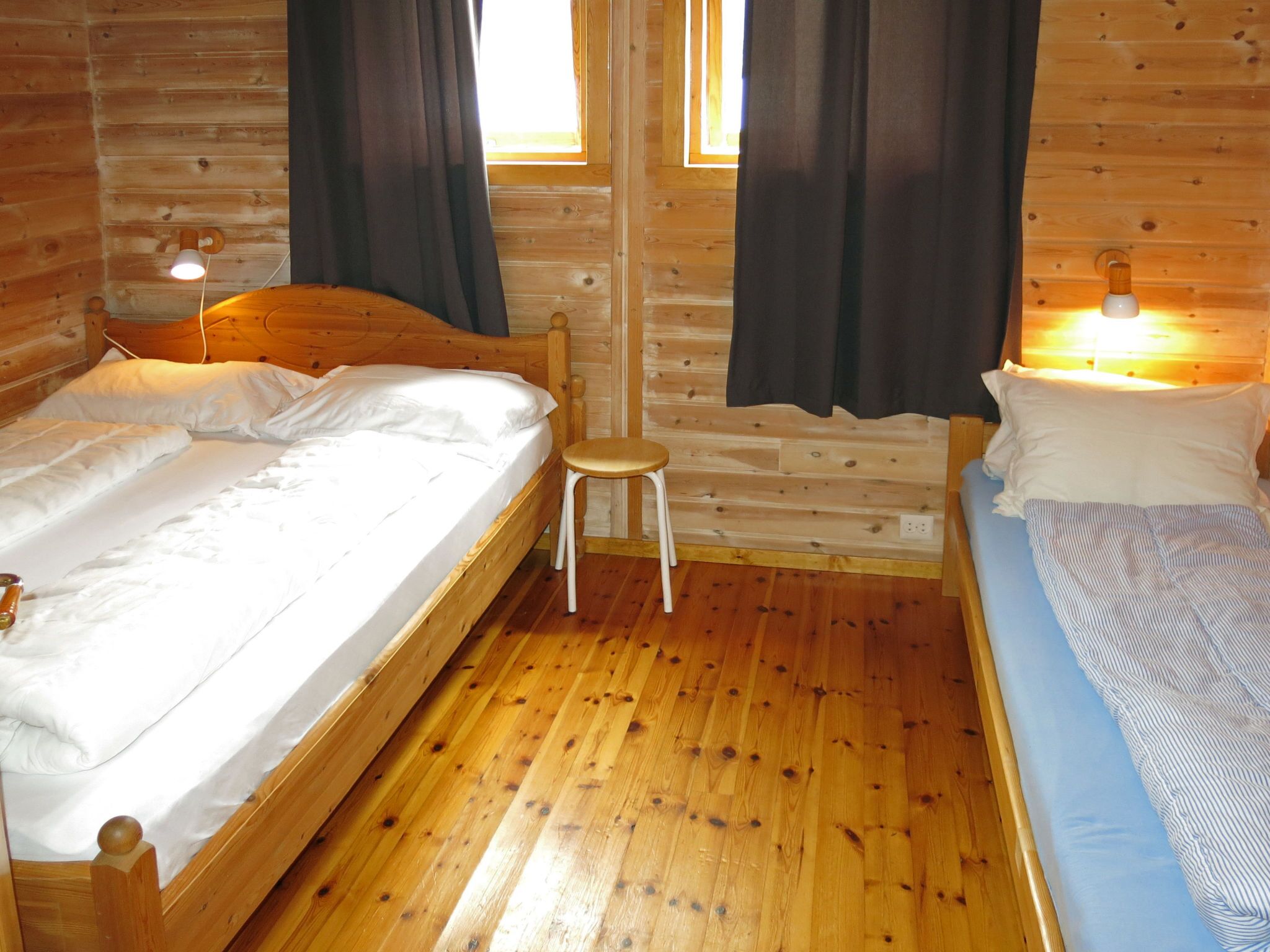 A wooden cabin room with two beds, a small stool, and a window with curtains.