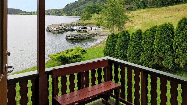 A wooden bench on a balcony overlooking a lake with boats, a dock, and a forested hillside. - Vacation home Nanna by Interhome (Risnes)