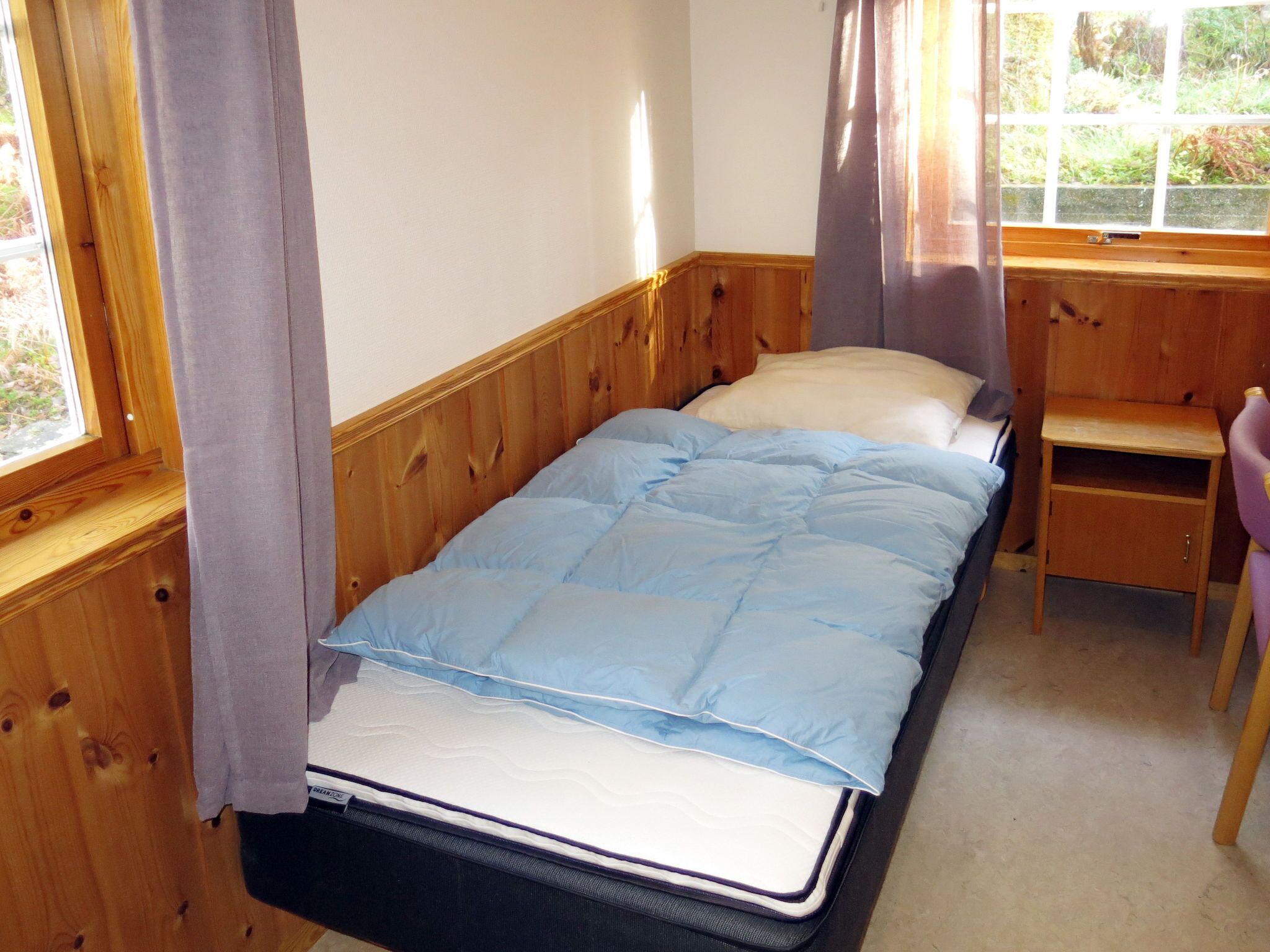 Ein Einzelbett mit blauer Matratze und Kissen, ein Holz-Kopfstück, ein kleiner Holztisch und ein Stuhl.