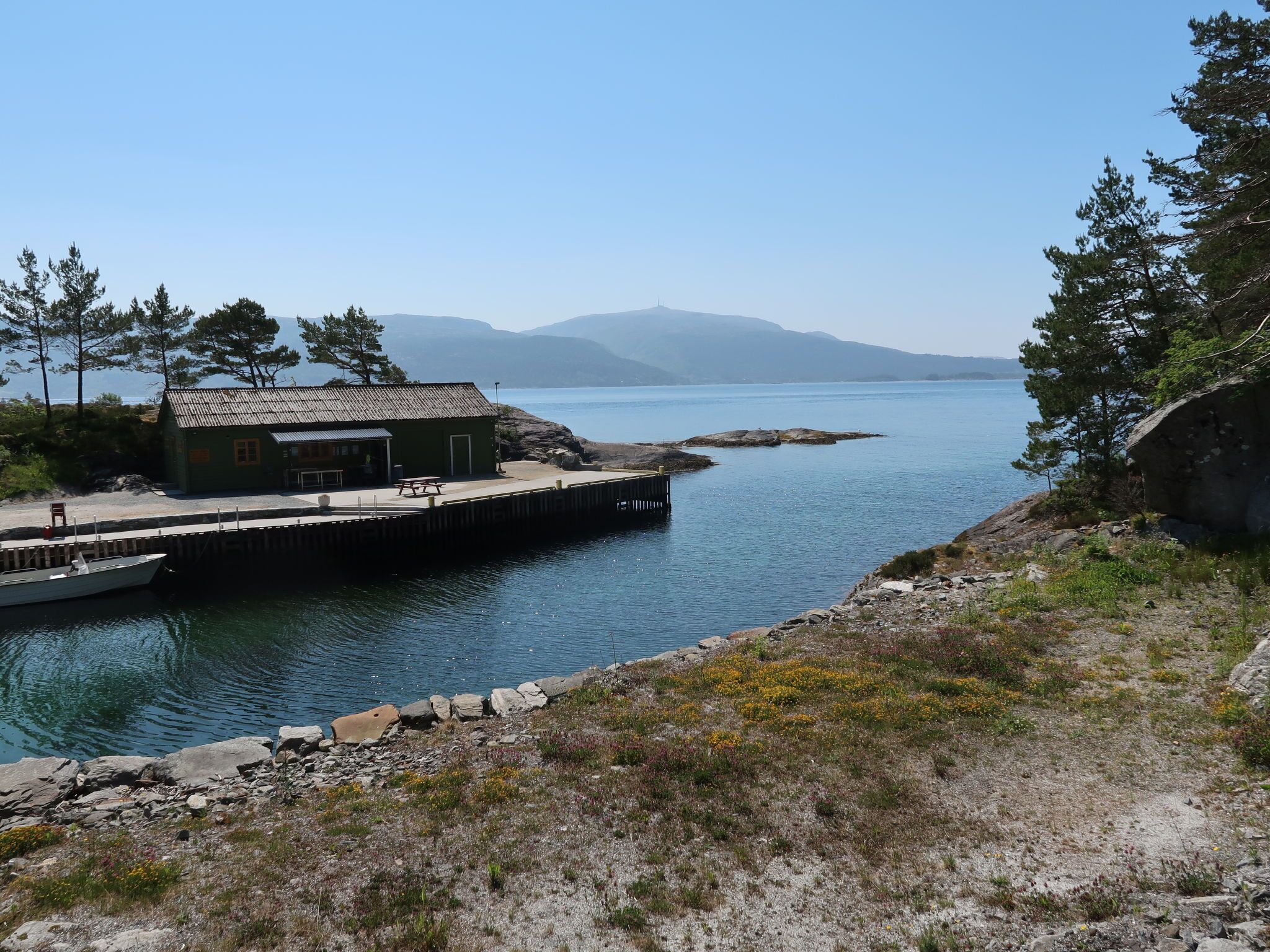 A small cabin with a dock extending into a calm lake, surrounded by rocky terrain and sparse vegetation.