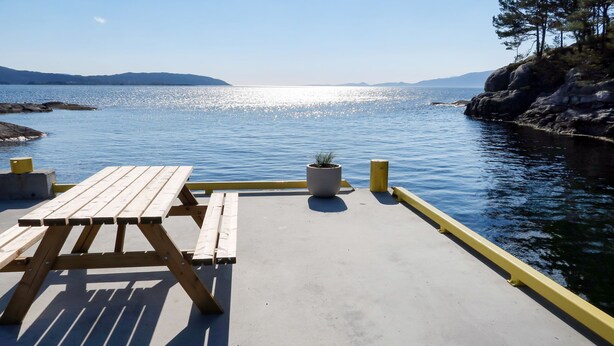 Una mesa de picnic de madera y un banco en un muelle de concreto, con una planta en maceta, con vistas a una masa de agua tranquila y colinas a la distancia.
