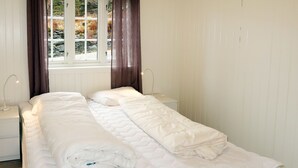 Cama doble con ropa de cama blanca, dos mesitas de noche con lámparas, una ventana con cortinas y vistas a árboles y rocas.