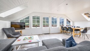 Een ruime woonkamer met een hoge plafond, een televisie, grijze banken, een eethoek met een houten tafel en stoelen, en grote ramen met uitzicht op de omgeving.