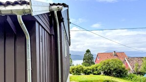 Ein Haus mit dunkler Fassade, einem sichtbaren Regenfallrohr und einem Rasen, der zu einem Haus mit rotem Dach im Hintergrund führt.