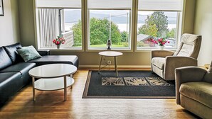 Ein Wohnzimmer mit einer Couch, zwei Sesseln, einem Couchtisch und einem Teppich. Drei Fenster mit Jalousien, eine Lampe auf einem kleinen Tisch und Blick auf Grünflächen und ein Gebäude.