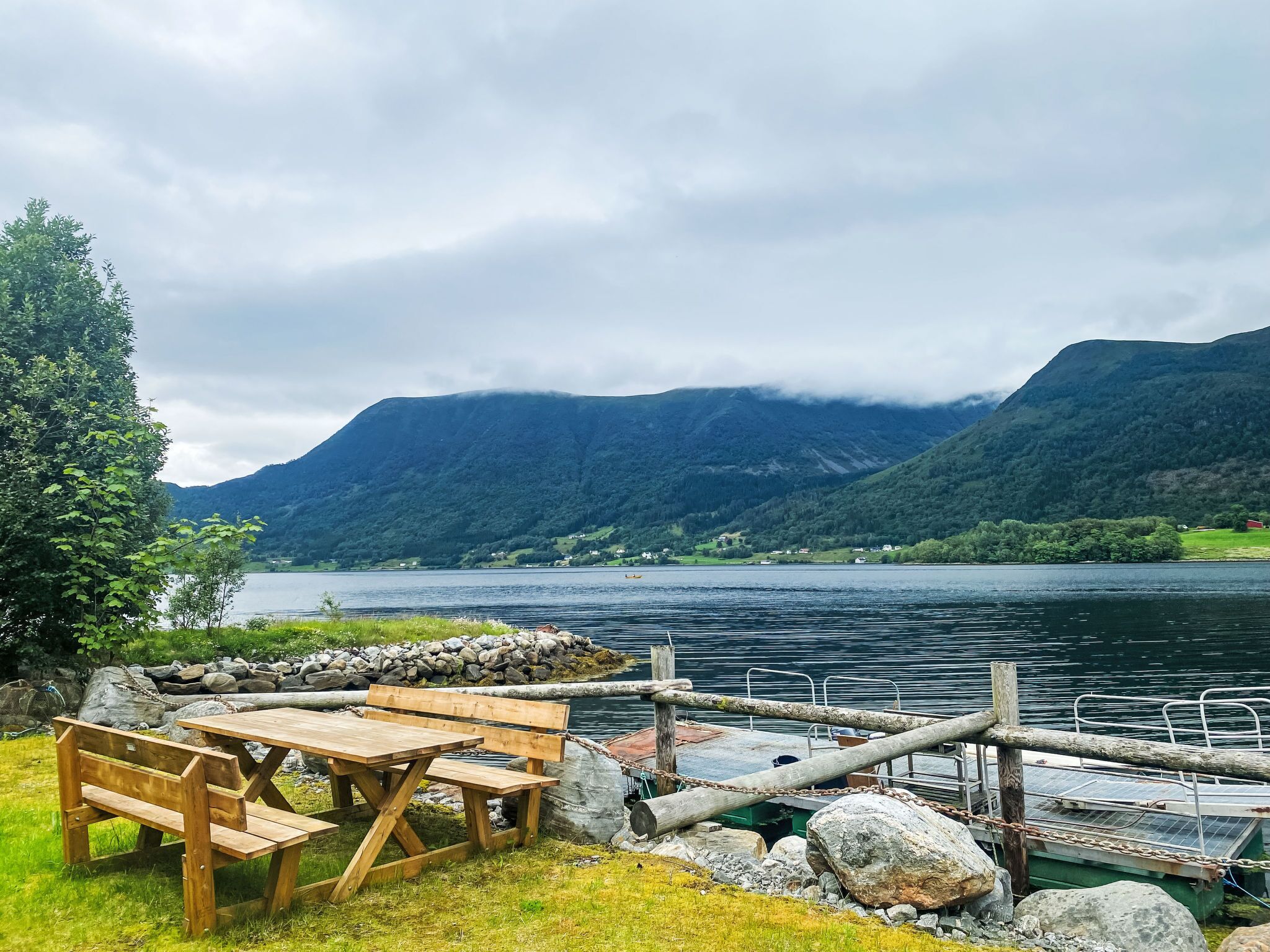 A lakeside picnic area with a wooden bench and table, surrounded by rocks and a dock.