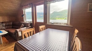 A cozy cabin interior with a wooden ceiling, a checkered table, a sofa, and two windows offering a view of a lake and mountains. - Naustet by Interhome (Åheim)
