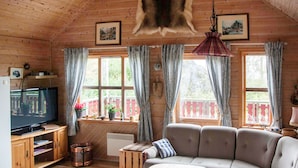 Interior acogedor de una cabaña con techo de madera, un televisor sobre un mueble de madera, un sofá, una mesita y una ventana con cortinas.