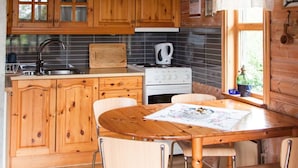 Interior de una cabaña de madera con cocina, que incluye armarios de madera, una estufa y una zona de comedor con mesa y sillas.