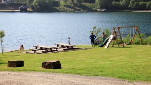 Un parc al costat d'un llac amb taules de pícnic, un parc infantil i cases al fons.
