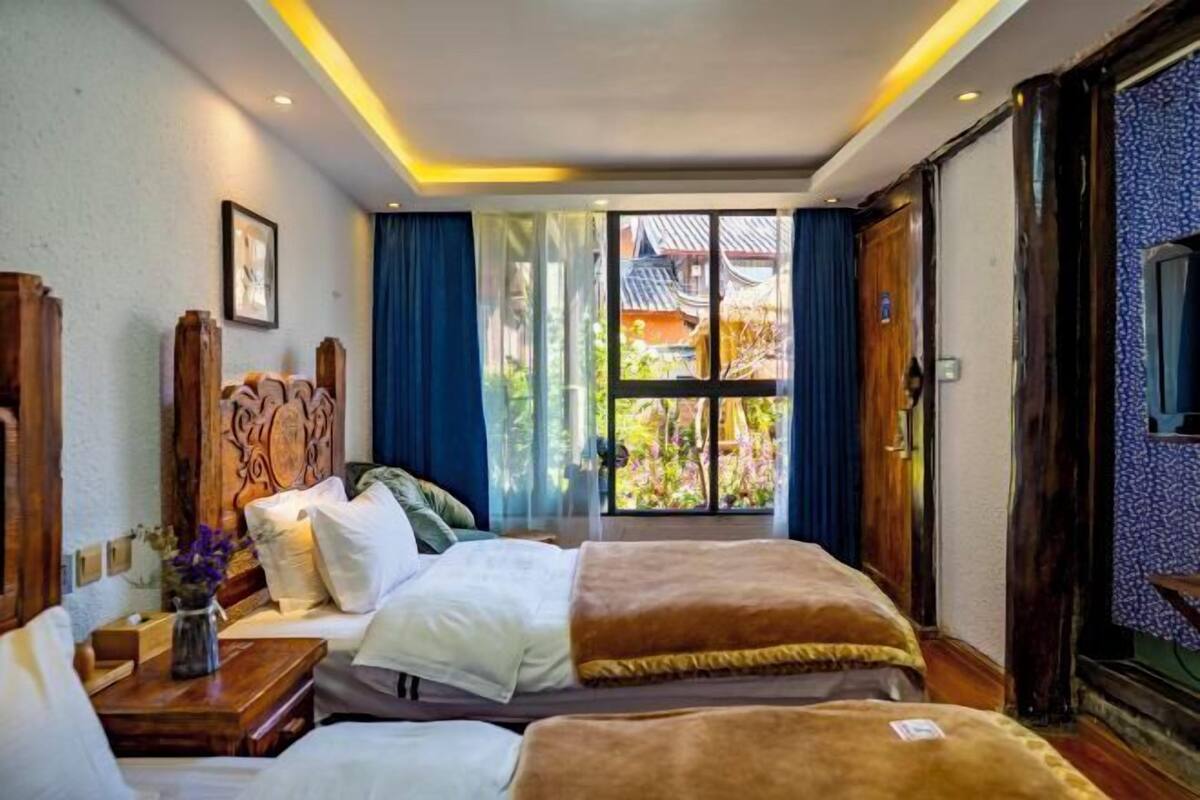 A hotel room with two beds, a wooden headboard, a desk with a vase of flowers, and a view of a traditional building through the window.