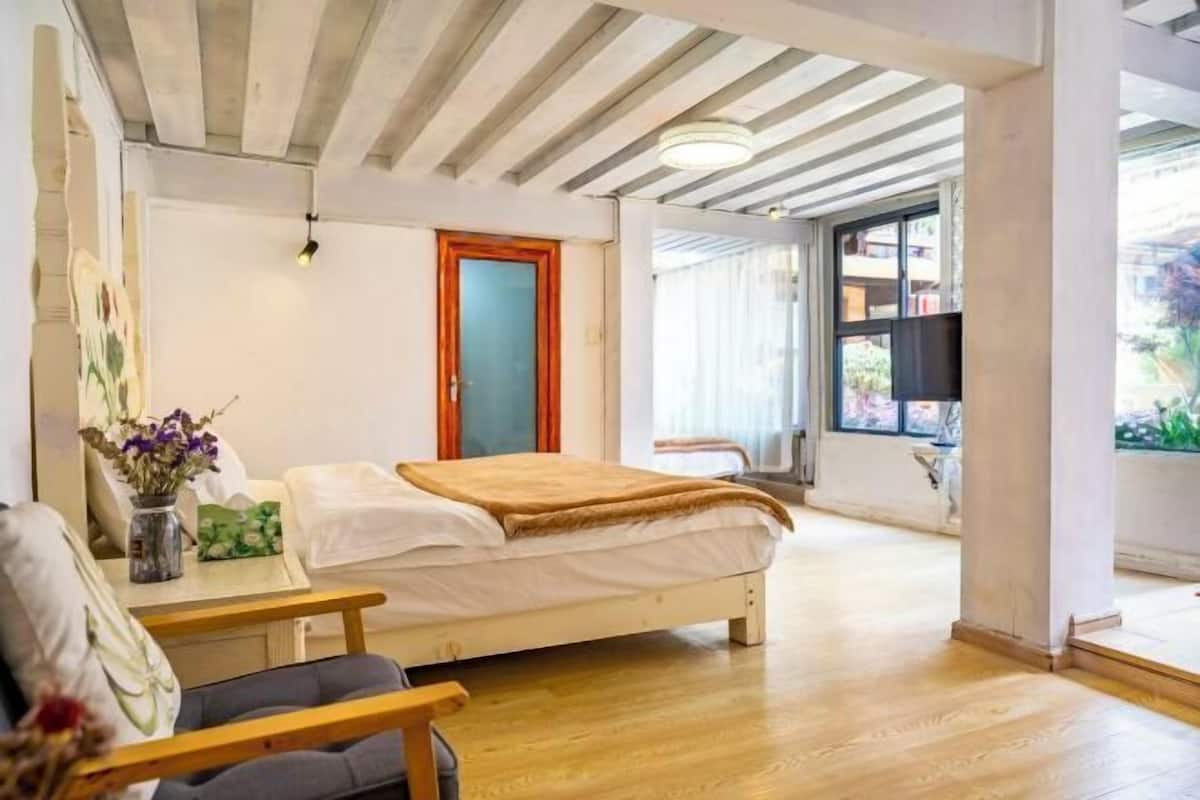 A bedroom with a bed, a bedside table with a vase of flowers, a chair, and a window with a view of the outside.